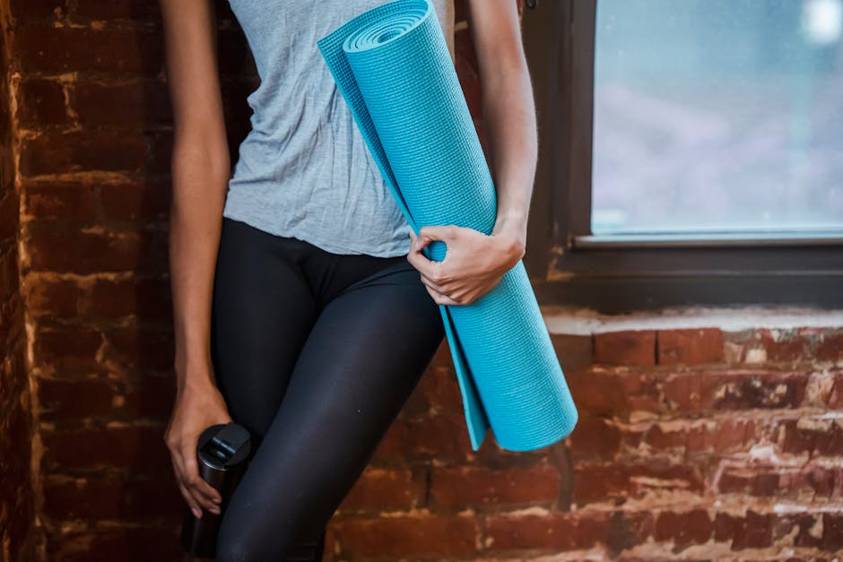 Unrecognizable sportive female in activewear standing near brick wall and window with rolled mat in hand during fitness training in gym