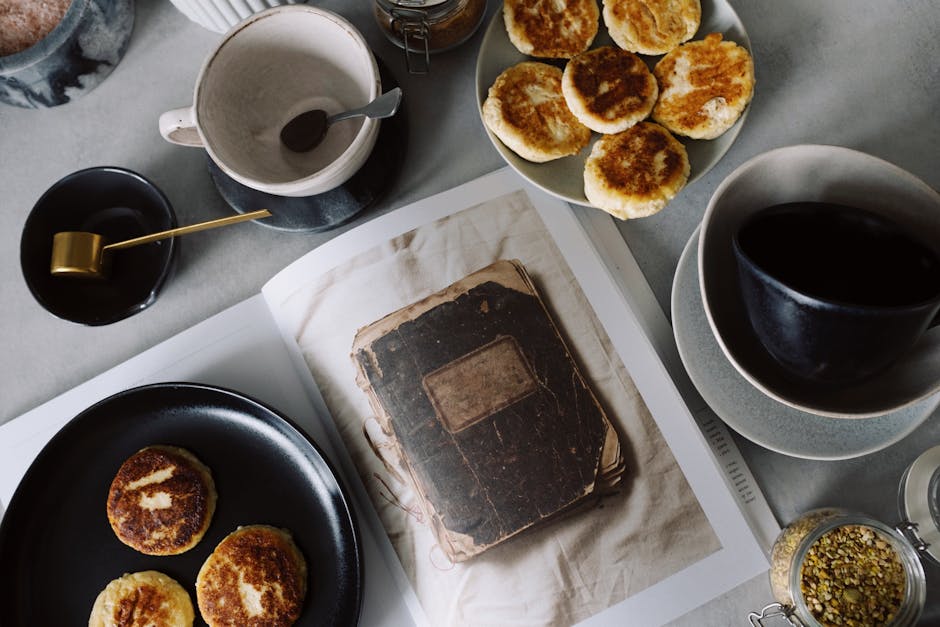 Top view of plate with delicious curd pancakes placed on recipe book surrounded by various plates on gray tabletop in kitchen