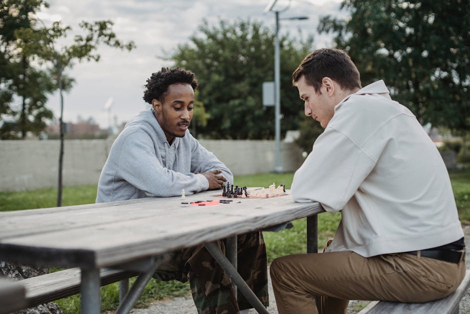 Side view of serious young guy playing chess with African American friends while spending time in city park