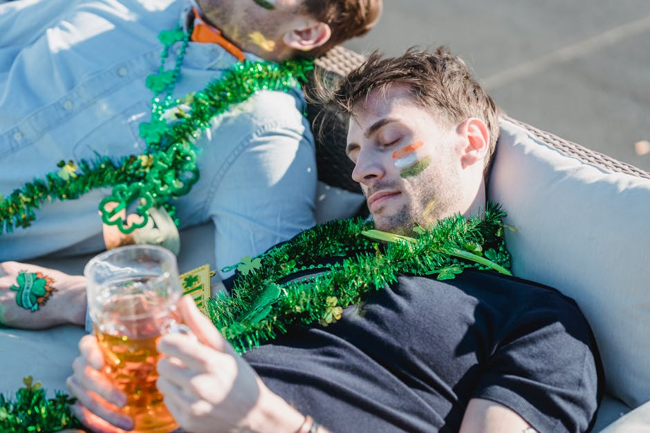 From above of drunk guy with glass of beer in hands lying on sofa and napping near crop unrecognizable friends after celebrating Feast of Saint Patrick