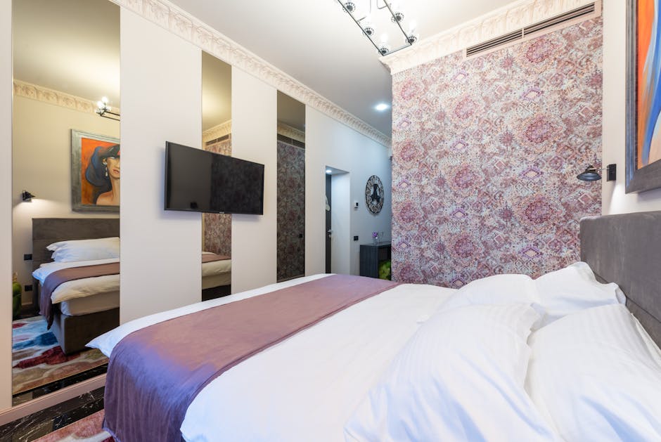 Stylish cozy light hotel suite with bed against TV hanging on wall with mirrors and wall with pink floral ornament