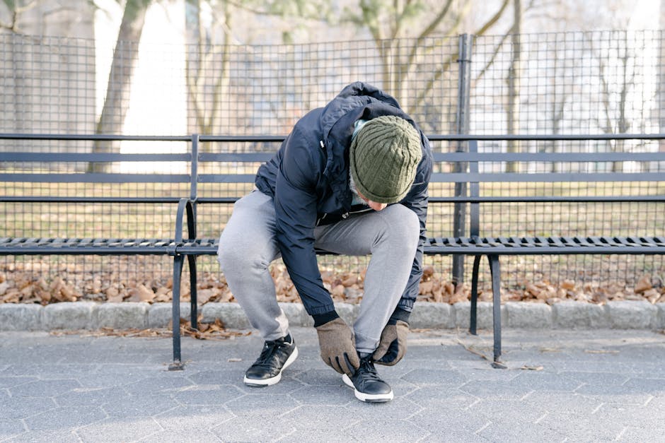 Full body of man in jacket with cap and gloves bending on bench in park while tying lace on sneaker