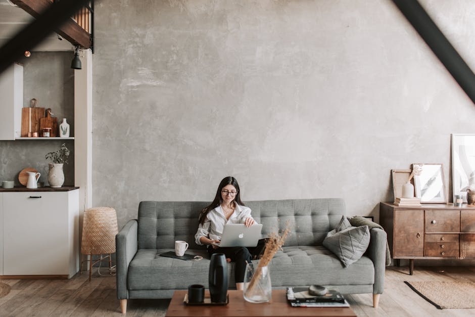 Happy female remote worker in eyeglasses sitting on cozy sofa with netbook and cup of coffee in loft style room with wooden floor and concrete wall