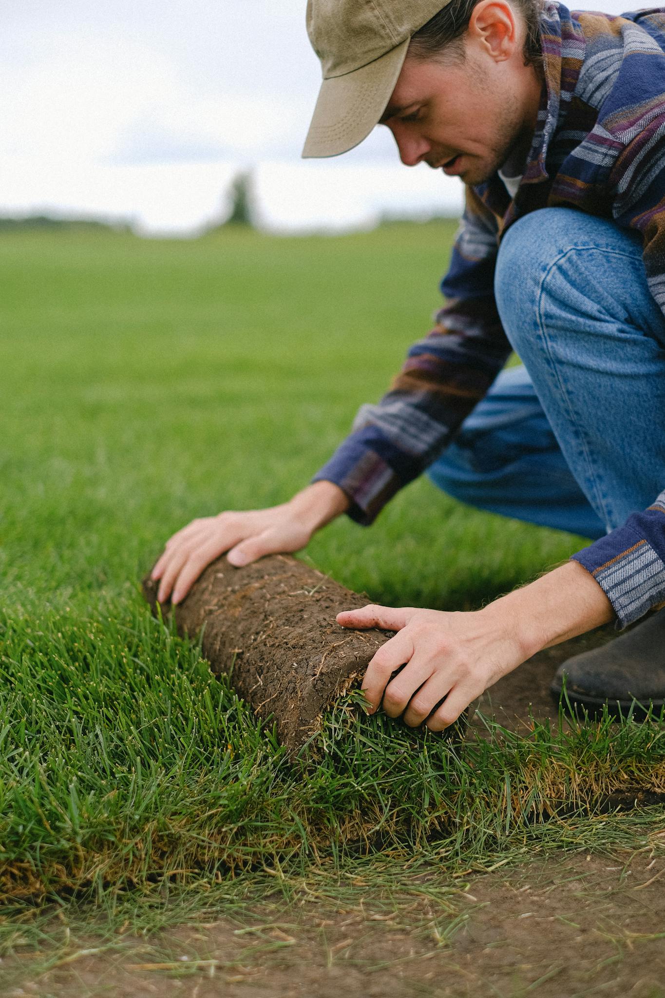 Side view of male worker laying sod grass onto ground for new garden lawn while working in countryside on blurred background