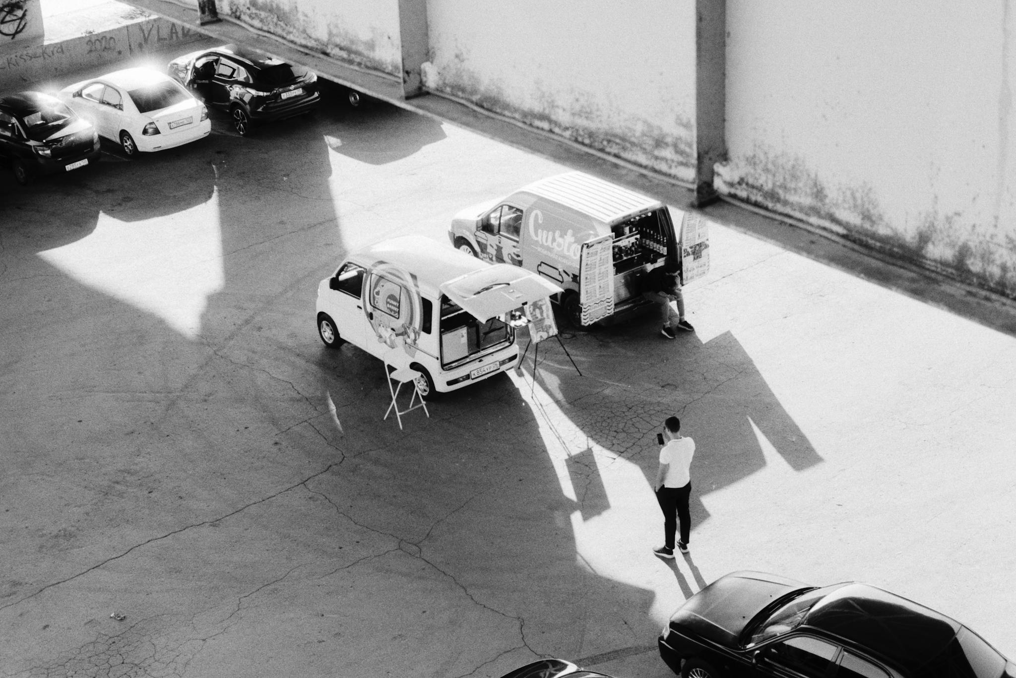 Black and white from above of man standing near coffee truck and car with easel on parking lot with cars