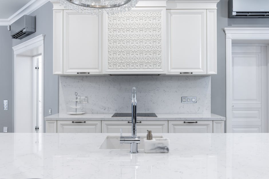 Interior of white classic kitchen with marble counter and chrome faucet in front of white cabinets next to door