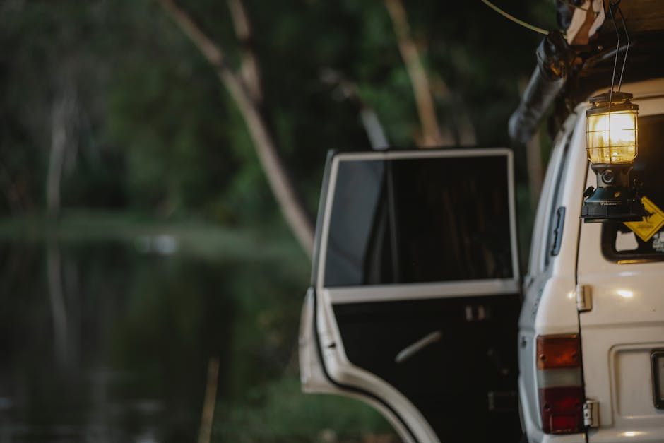 Glowing vintage lamp hanging from SUV car with opened door parked on lake shore during camping in green forest in evening