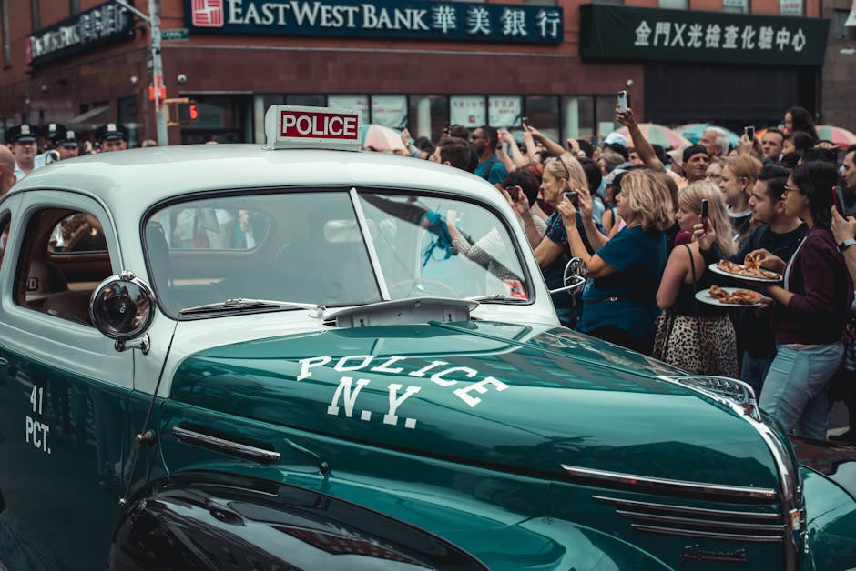 A Crowd Of People Gathered Around A Green Vintage Police Car In The City Street