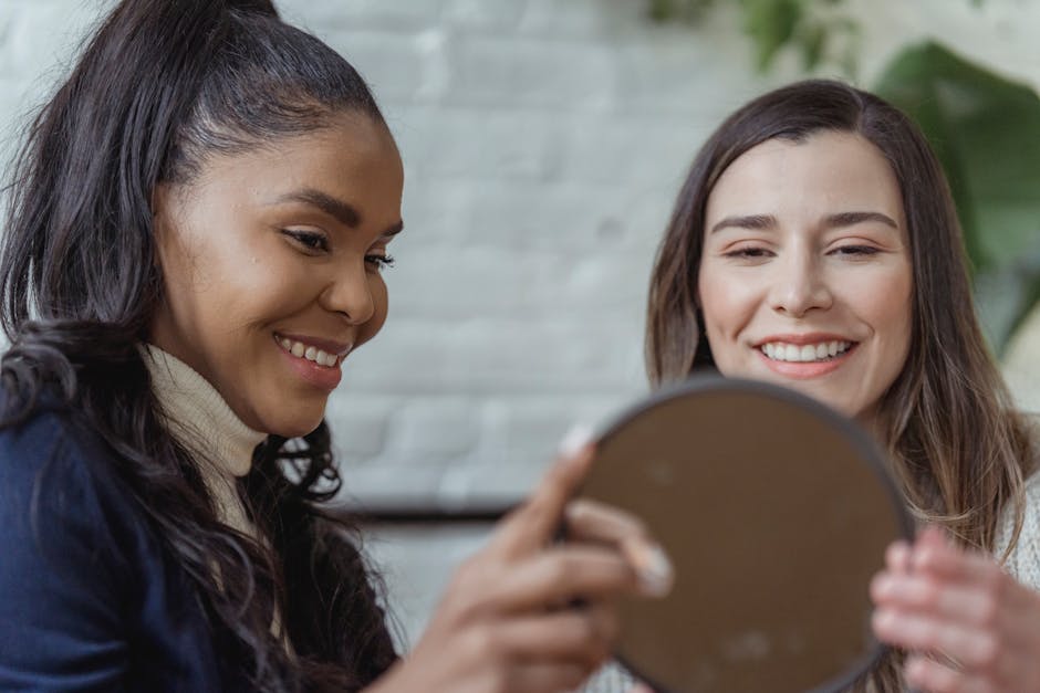 Happy young female client looking in mirror and smiling near positive ethnic makeup artist