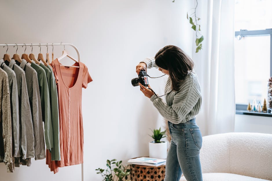 Side view of young female photographer in casual outfit standing in front of hanger and taking picture with camera of shirt in modern apartment