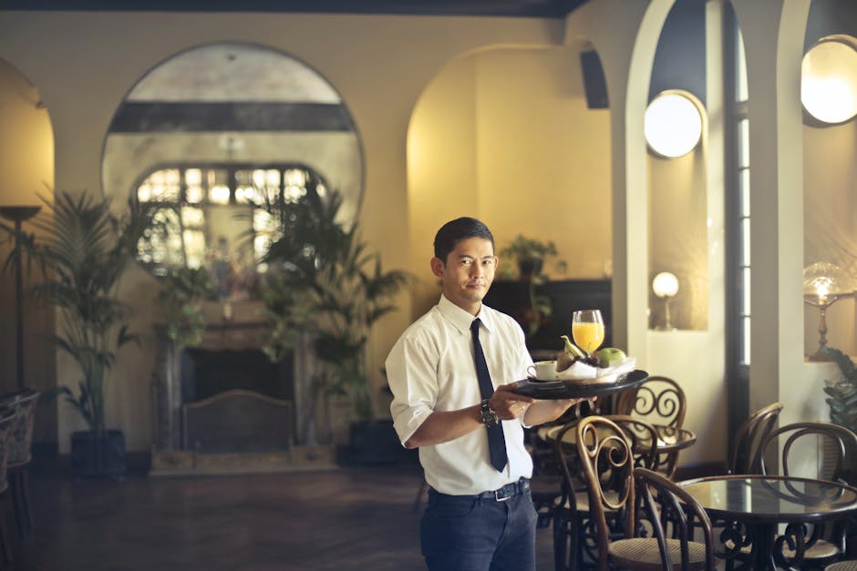 Confident young ethnic waiter in elegant clothes holding tray with food and drinks and looking at camera while serving tables in stylish restaurant