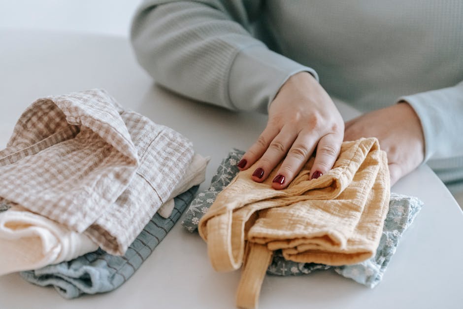 Crop faceless female with red manicure folding colorful baby garments while sitting at white table in light room at home