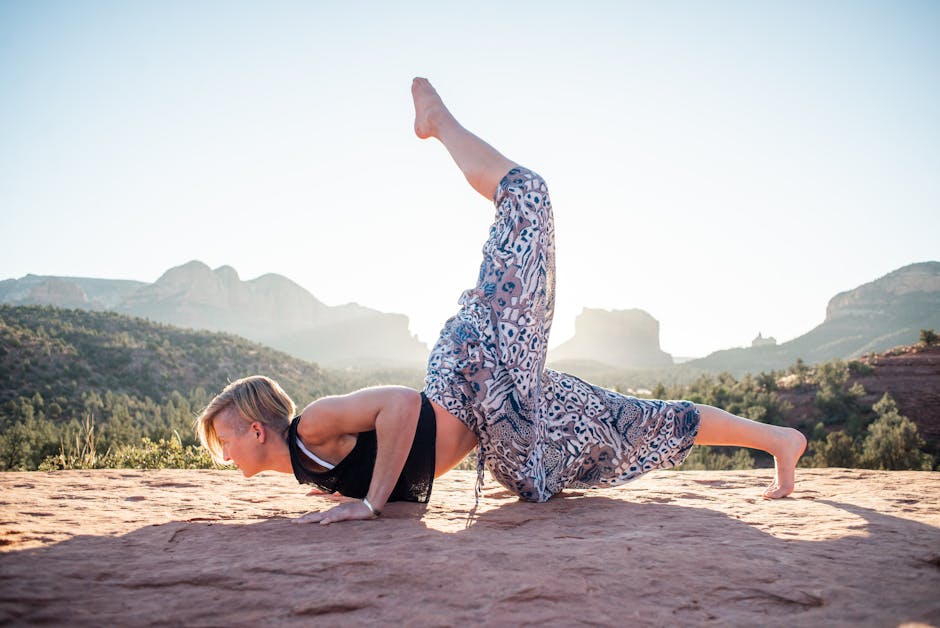 Side view of short haired female in summer clothes doing Ganda Bherundasana while practising yoga on sunlit cliff with mountains in background