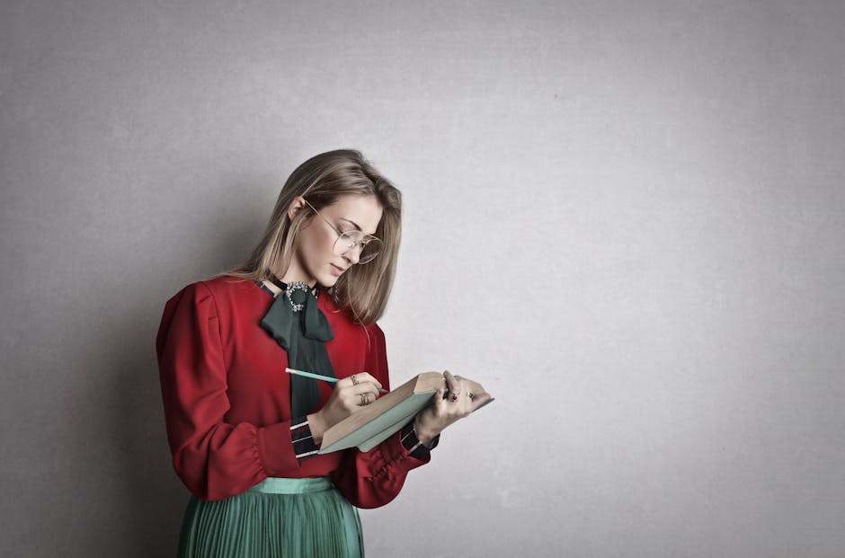 Pensive attentive woman in glasses and elegant vintage outfit focusing and taking notes with pencil in book while standing against gray wall