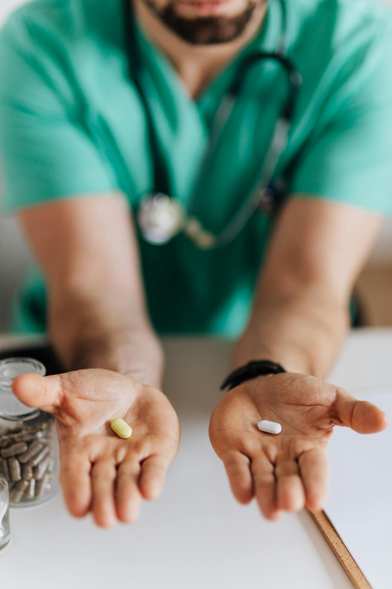 Crop man doctor wearing medical uniform demonstrating medicine to patient in clinic during work sitting at table with stethoscope and clipboard