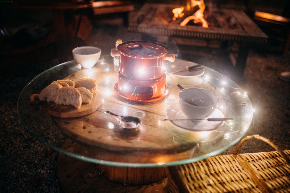 Small fondue pot and skewers with cups placed on woodn and glass table with served bread pieces for picnic in night garden