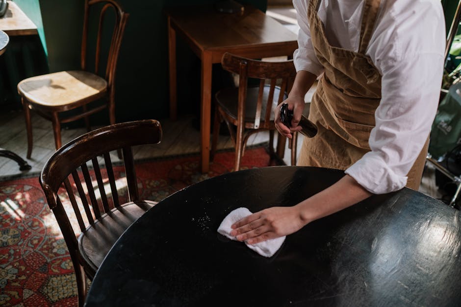 Person in White Shirt and Brown Apron Cleaning Black Table with White Wipe