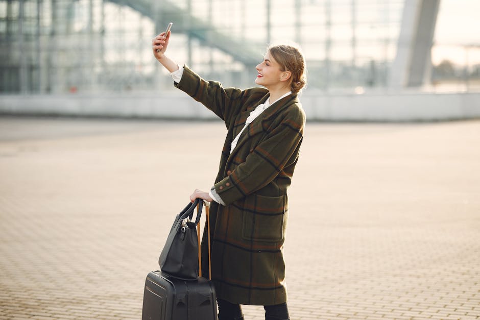 Stylish young woman with luggage taking selfie outside modern glass building