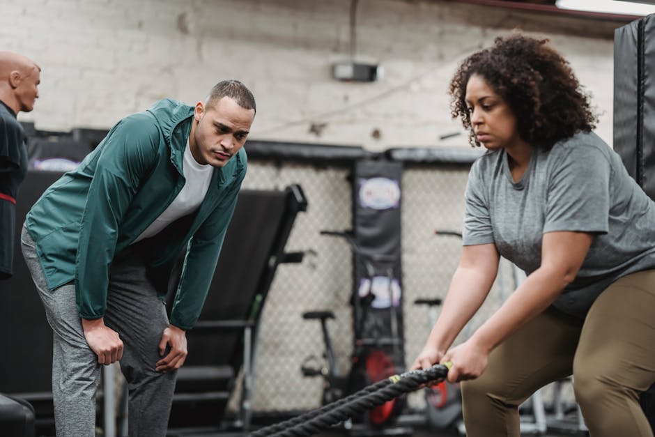 Pensive professional instructor supporting concentrated plump African American woman in training with gym equipment