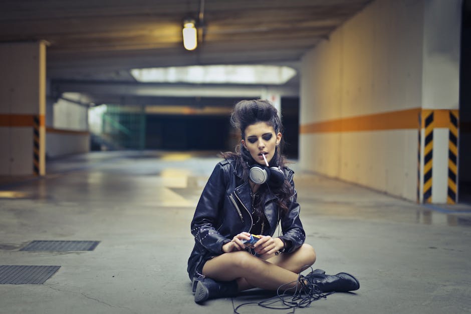 Young stylish woman wearing leather jacket and boots sitting with legs crossed on concrete ground with headphones and smoking cigarette