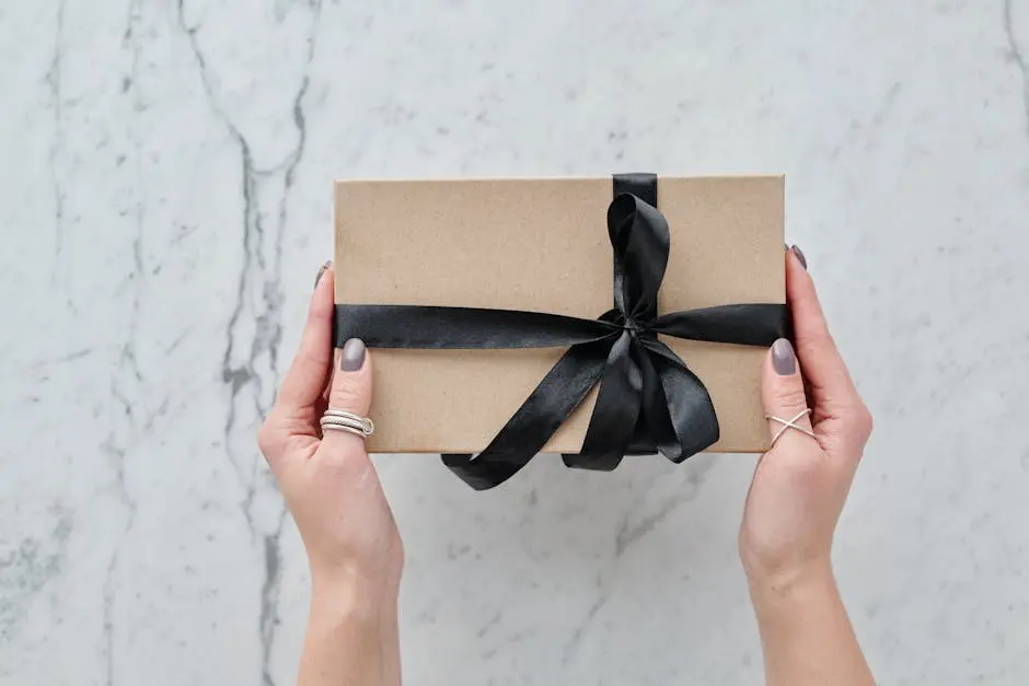 A beautifully wrapped brown gift box with a black ribbon held by two hands over a marble surface.