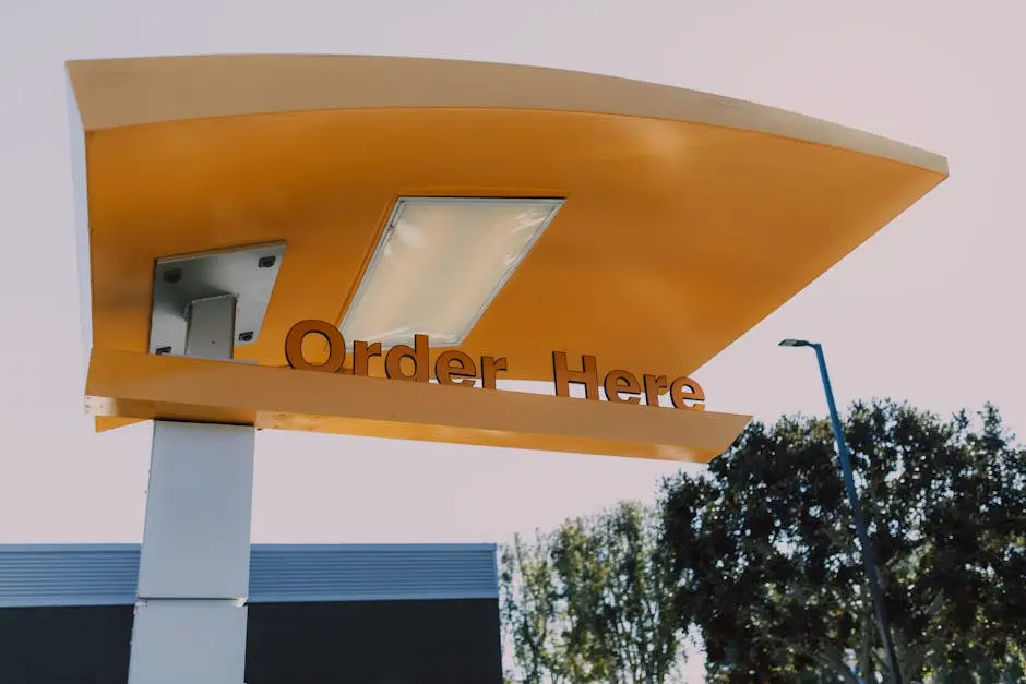 A modern drive-thru order sign with yellow canopy under a clear sky, surrounded by trees.