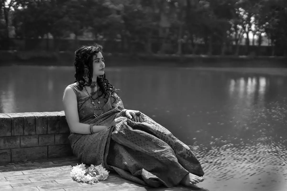 A woman in traditional attire sits gracefully by a tranquil river, exuding a sense of peace.