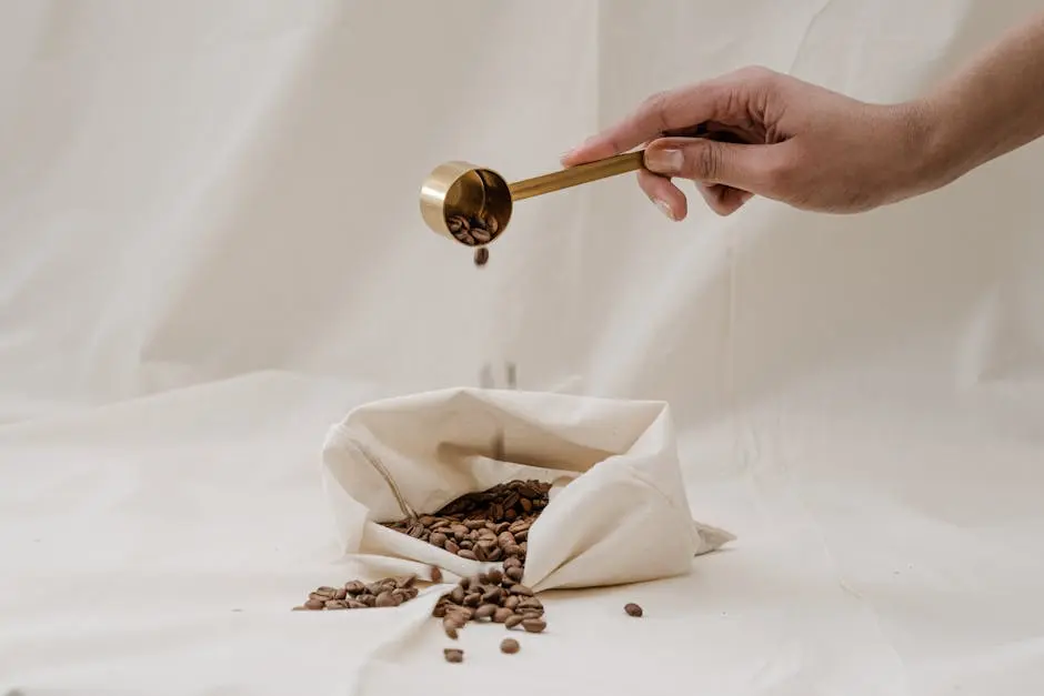 Hand pouring coffee beans with a gold scoop into an eco-friendly bag on a soft background.