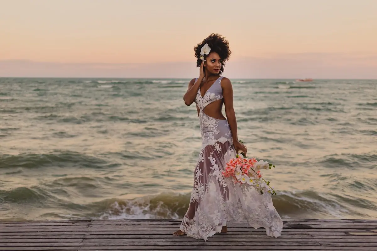 Stylish black bride with flower bouquet walking near ocean