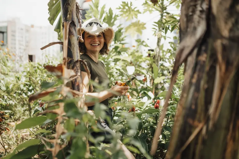 Smiling woman with a hat tending plants in an urban garden, surrounded by greenery on a bright summer day.