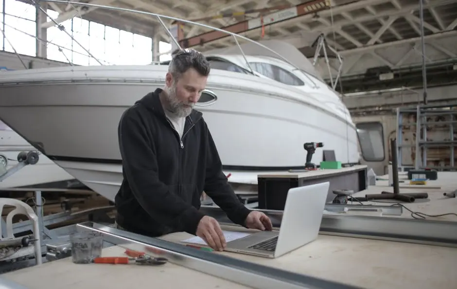 Mechanic using laptop in a boat workshop, focusing on yacht maintenance and repair tasks.