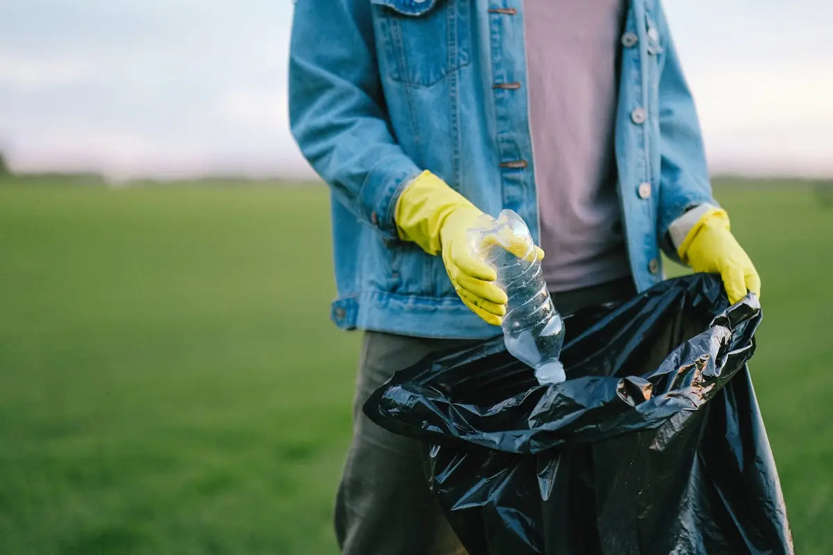 Person in Blue Denim Jacket Putting Plastic Bottle Inside Black Garbage Bag