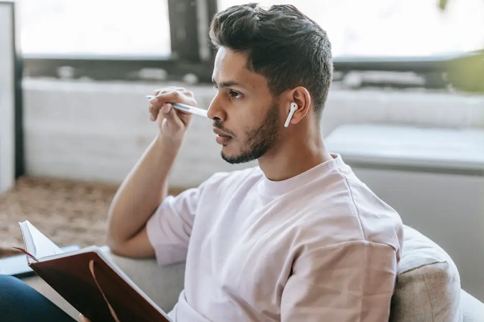 Concentrated young ethnic male in modern earbuds taking notes in planner while sitting in armchair and looking away in contemplation