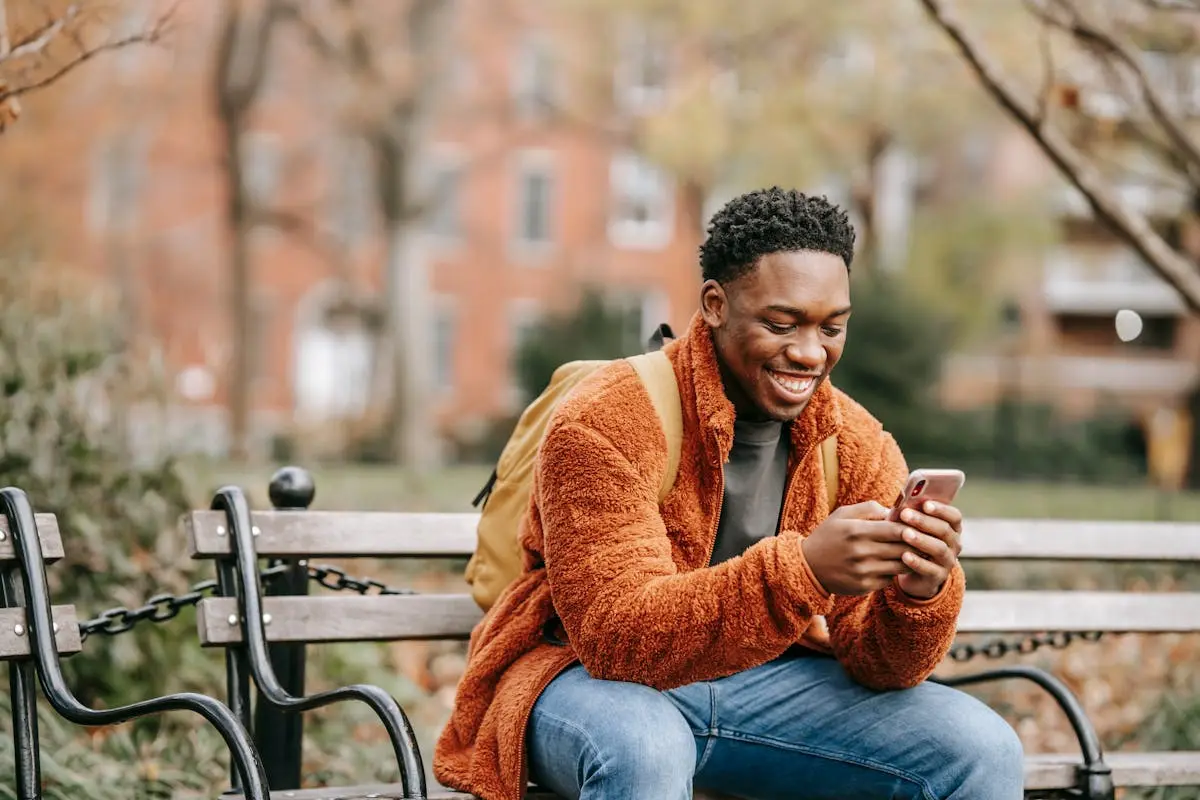 A cheerful young man in warm clothes smiling while using his phone on a bench in a city park.