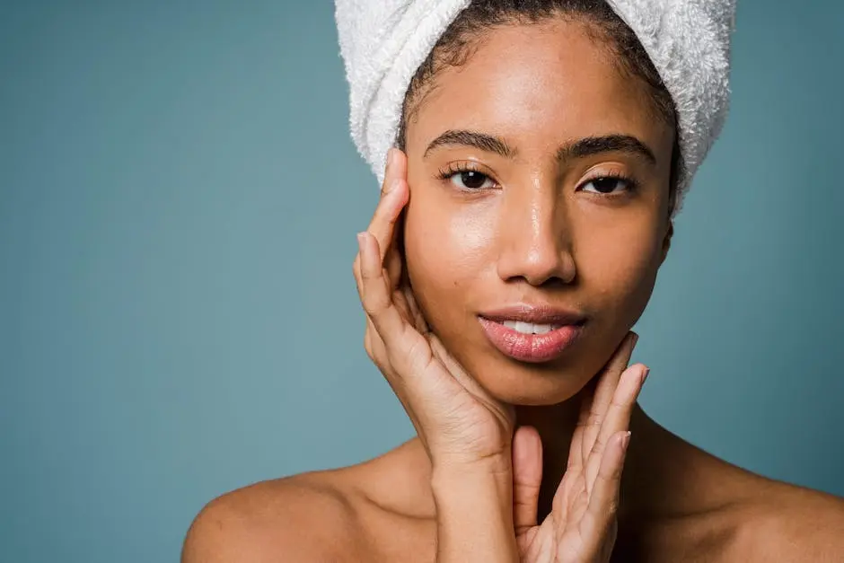 Calm African American female with towel turban on head and bare shoulders touching face and looking at camera against blue background in studio