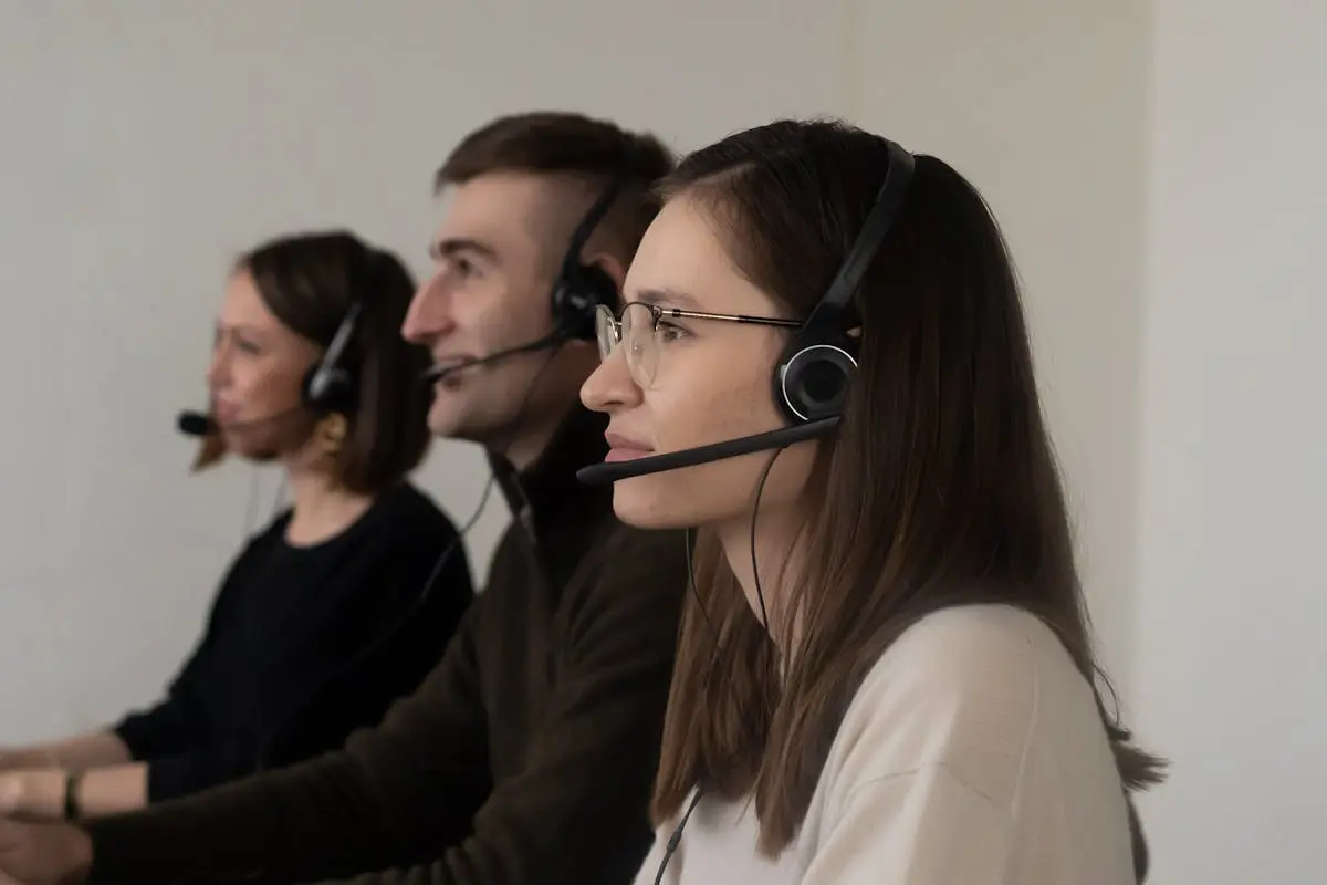 Side view of young positive operators in casual outfit with headphones with microphone in light workplace