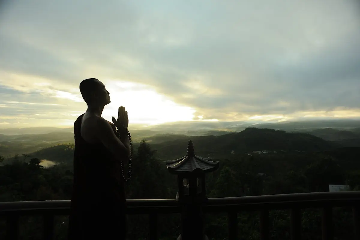 A monk in silhouette prays peacefully at sunrise with mountains in the background.