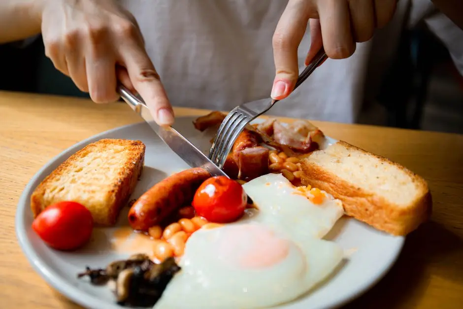 Hand slicing a full English breakfast with bacon, sausage, eggs, and tomatoes.