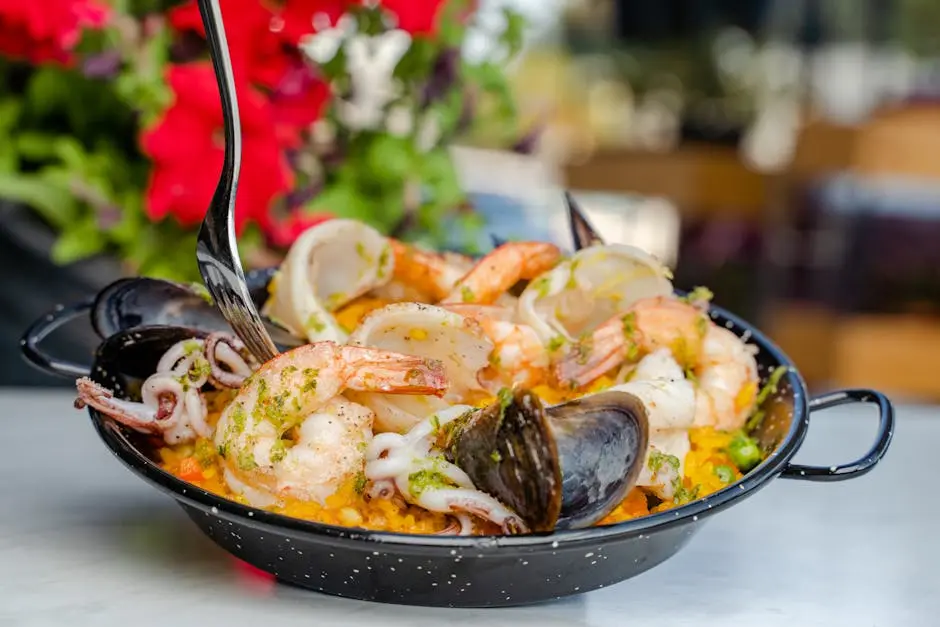 Close-up of a seafood paella dish in a black pan, garnished with shrimp, mussels, and calamari, served outdoors.