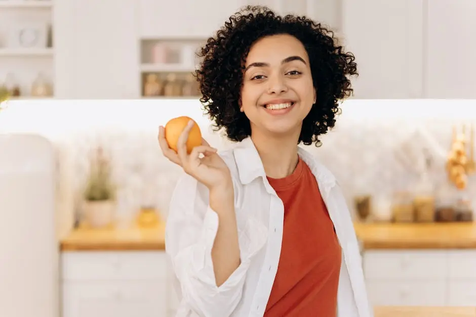 A joyful woman with curly hair holding an orange in her kitchen, promoting a healthy lifestyle.