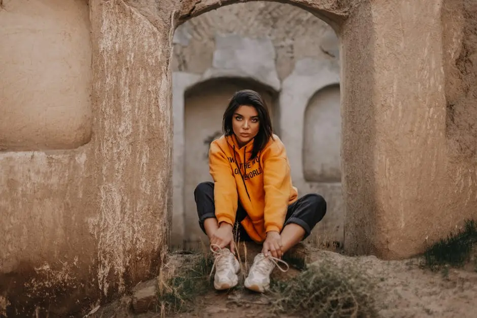 Stylish young woman in a vibrant orange hoodie and sneakers sitting outdoors in a rustic setting.