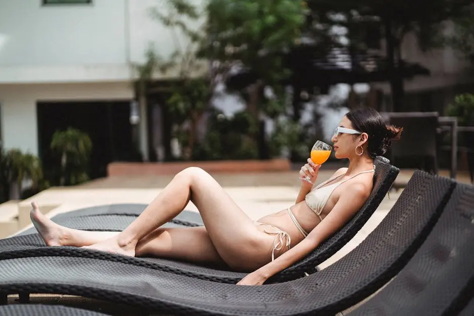 Side view of young ethnic barefooted lady in stylish swimwear and sunglasses drinking cold orange juice while sunbathing at poolside in modern hotel