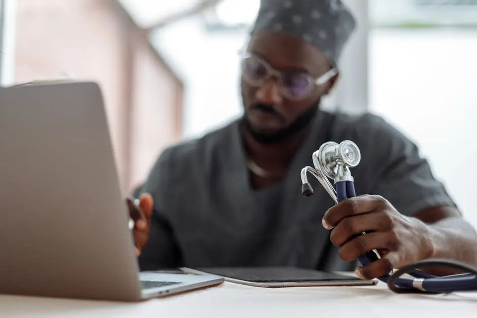 A Man in Gray Scrub Suit Holding Stethoscope while Typing on His Laptop