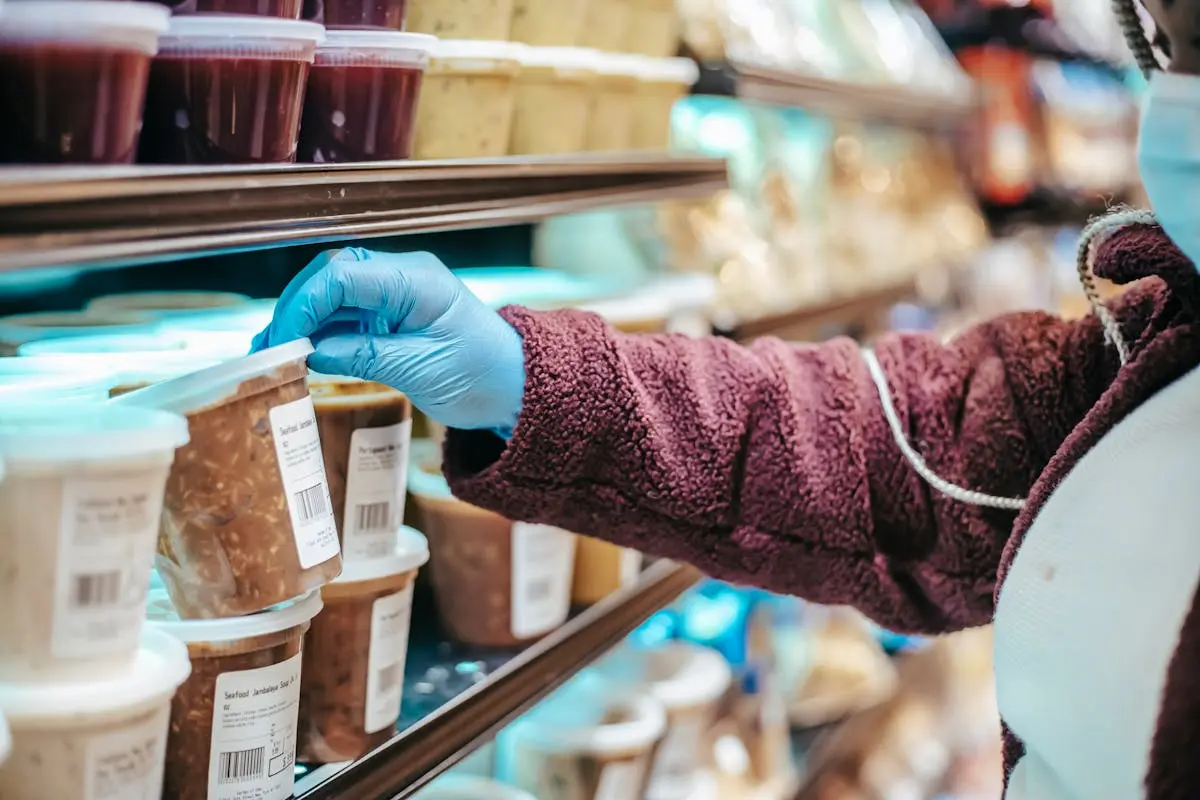 Crop anonymous female customer in protective mask reading label on frozen food in plastic container in grocery store