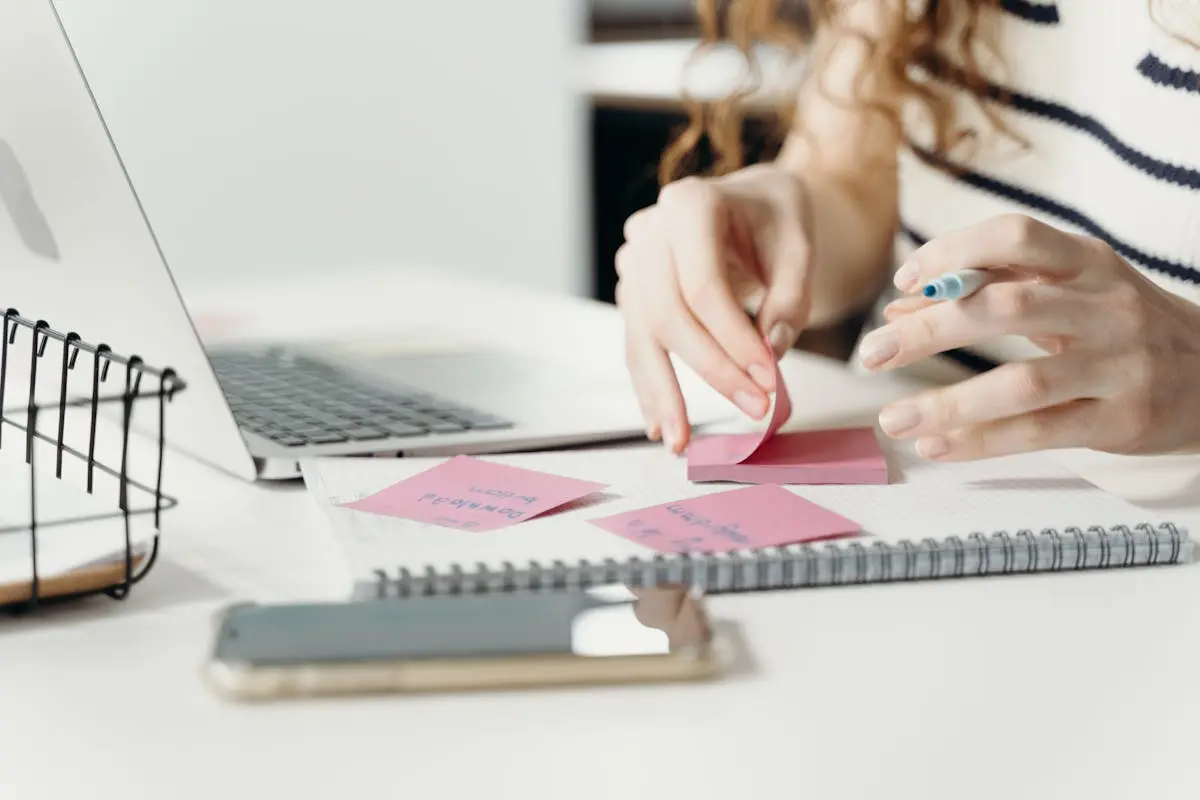 Woman organizing notes with a laptop and smartphone at a modern workplace.