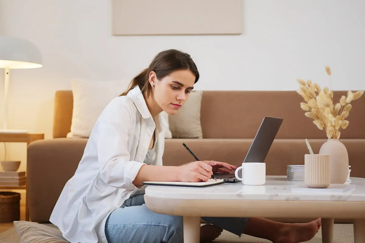 Side view full length young focused female in casual clothes browsing modern laptop and writing down notes while sitting on floor in cozy modern apartment