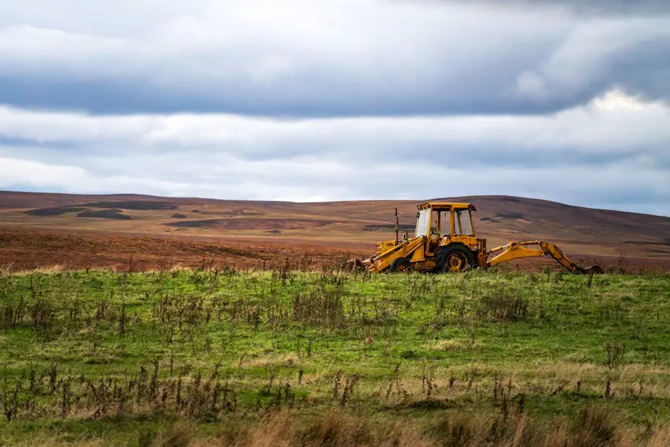 A solitary yellow backhoe on a wide green field against a backdrop of hills and a cloudy sky.