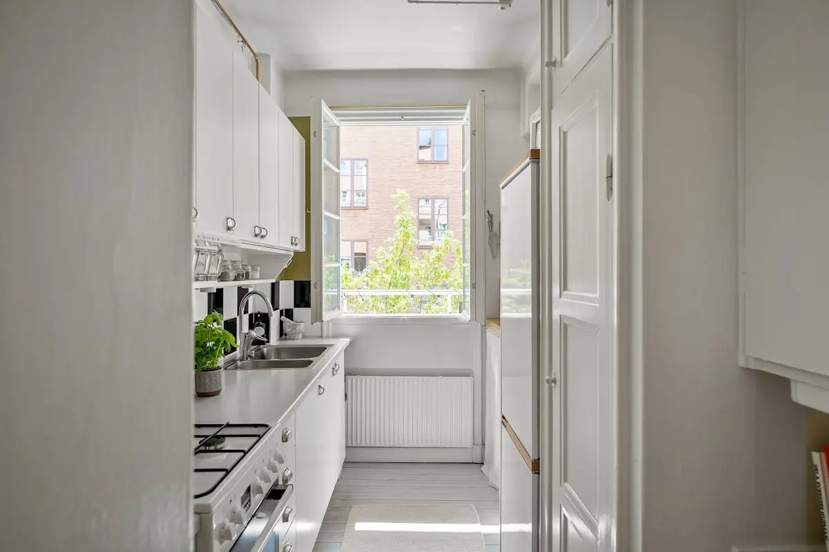 A narrow kitchen with white cabinets and a window