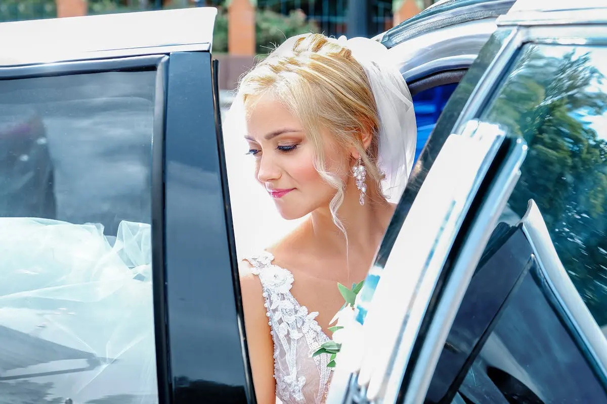 Charming young bride in white dress and veil sitting in black modern car on sunny summer day
