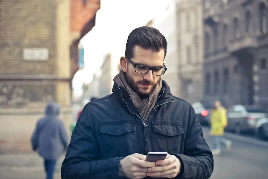 Man Wearing Black Zip Jacket Holding Smartphone Surrounded by Grey Concrete Buildings